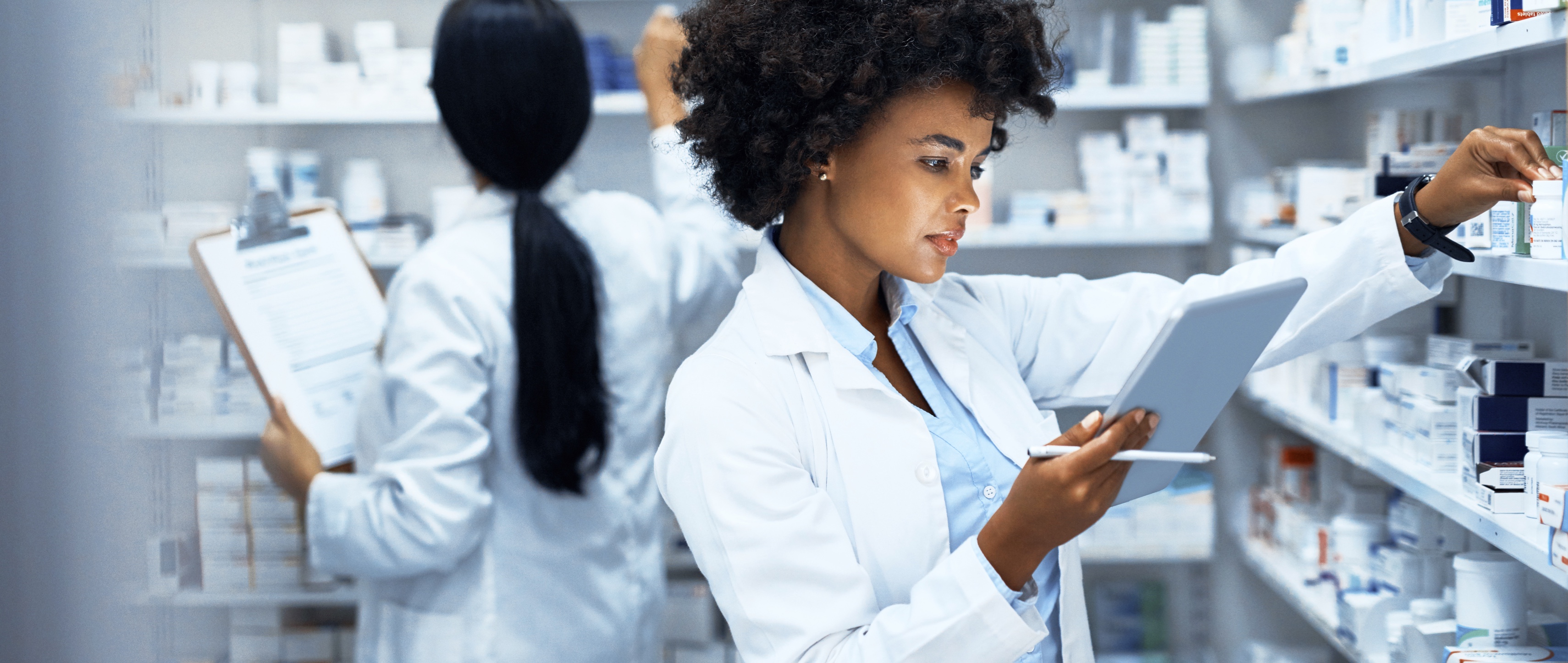 young woman doing inventory in a pharmacy on a digital tablet with her colleague in the background